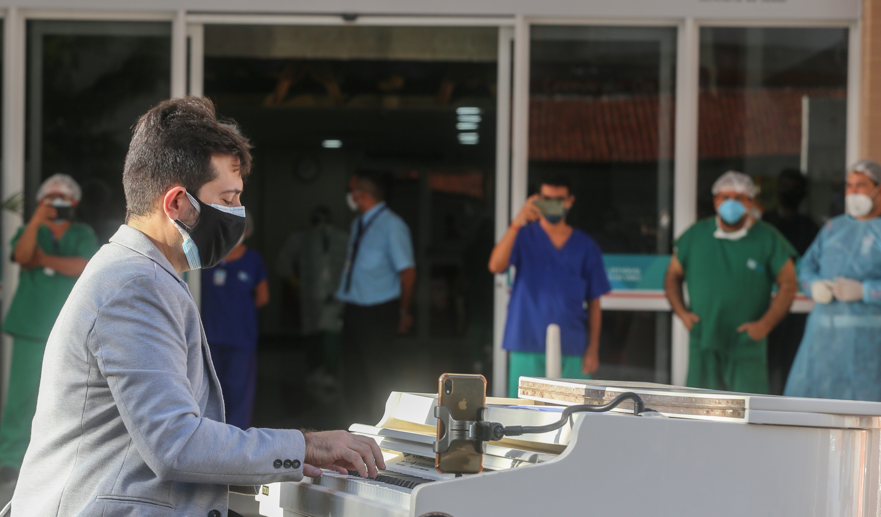 um pianista em cima de um caminhão tocando em frente a um hospital enquanto é observado por profissionais de saúde. Todos estão com máscara